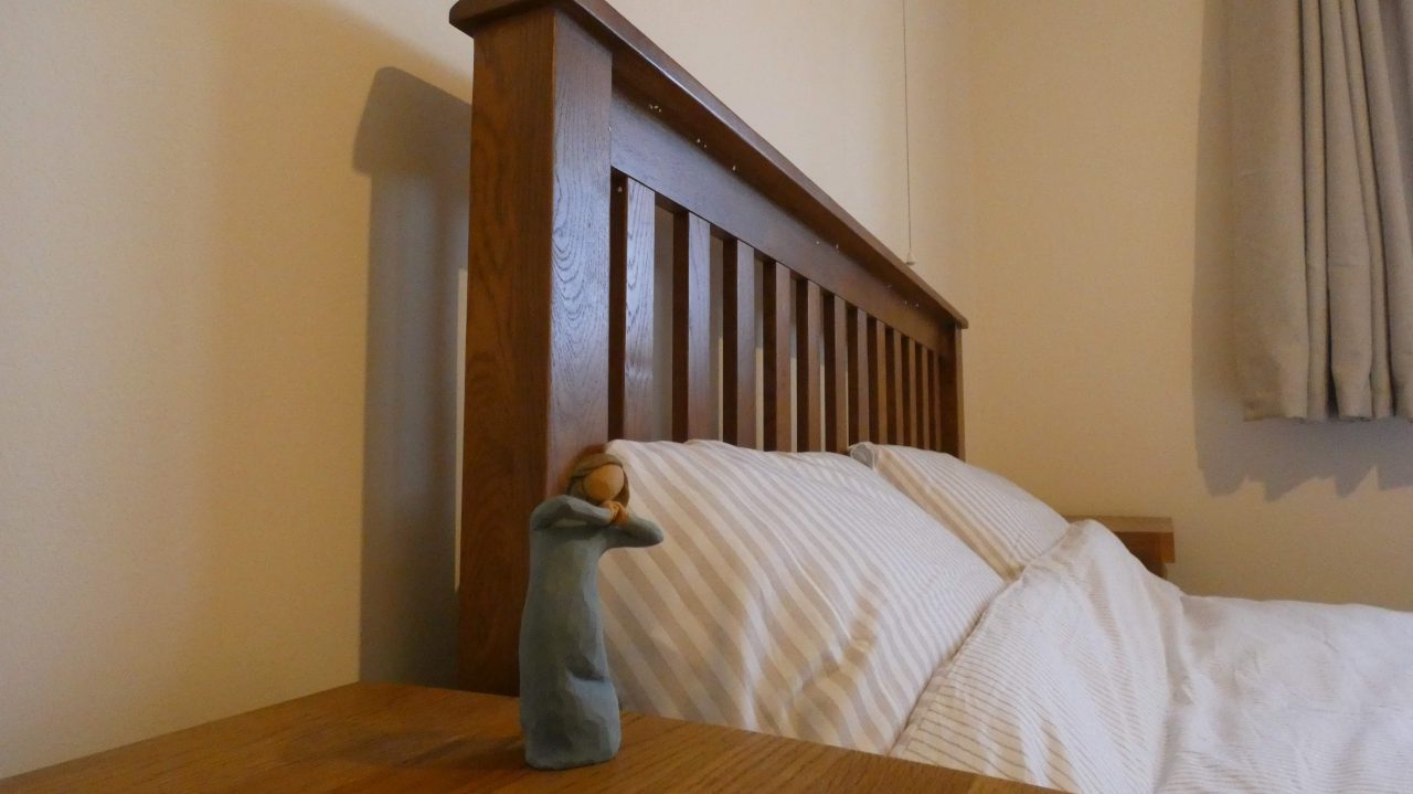 Bed against a cream wall. The headboard is made out of wood and the bedding is white. On the bedside table sits a small figurine.