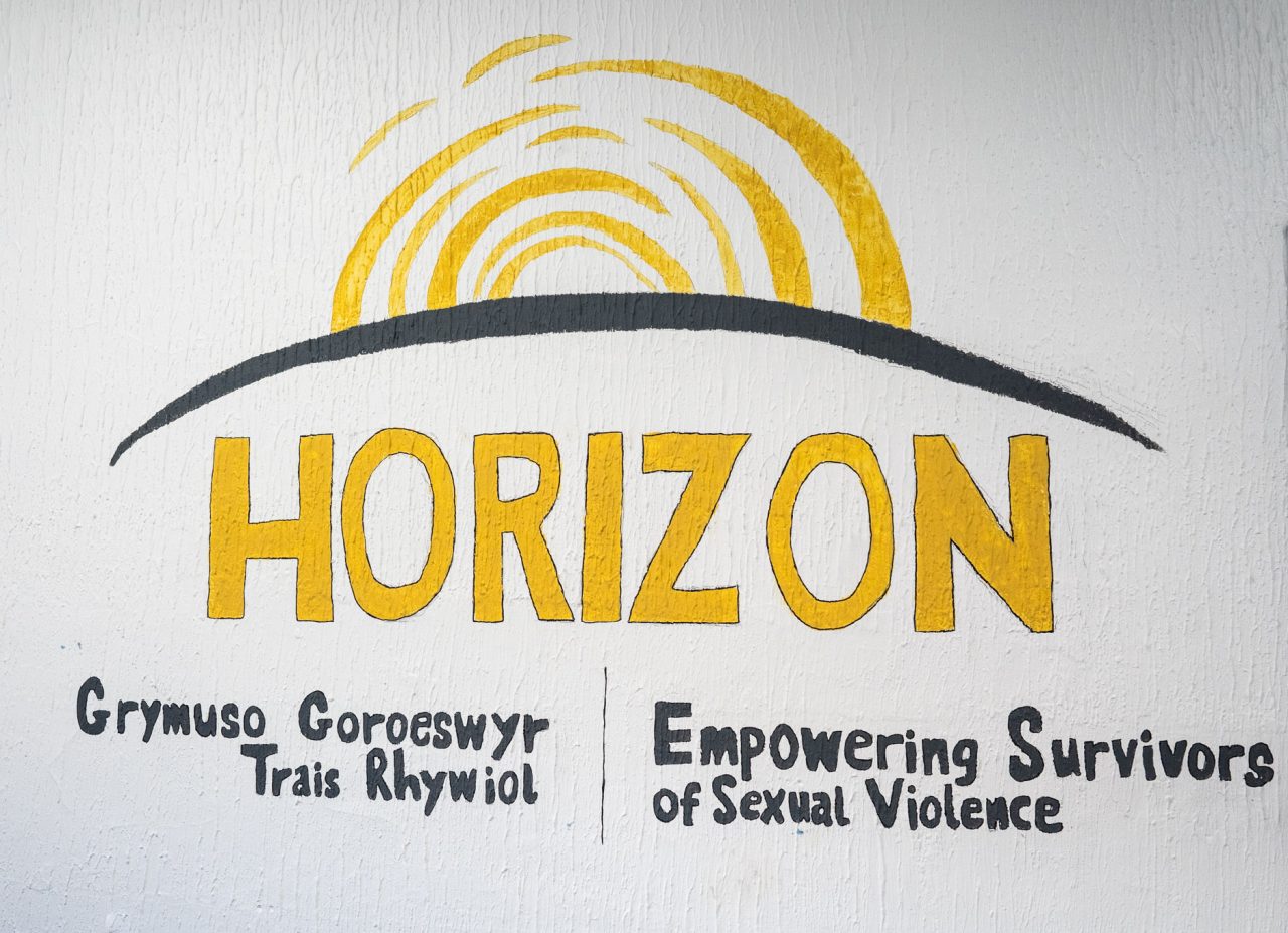 A drawing of the words 'horizon', which appear in yellow caps. Above it is a drawing of a yellow sunrise. Underneath is are the words, 'Empowering survivors of sexual violence' and 'Grymuso Goroeswyr Trais Rhywoil'.