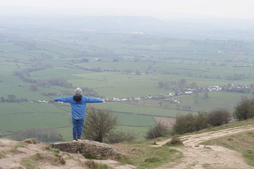 Person, with their back to the camera, standing on top of a mountain with their arms outstretched.