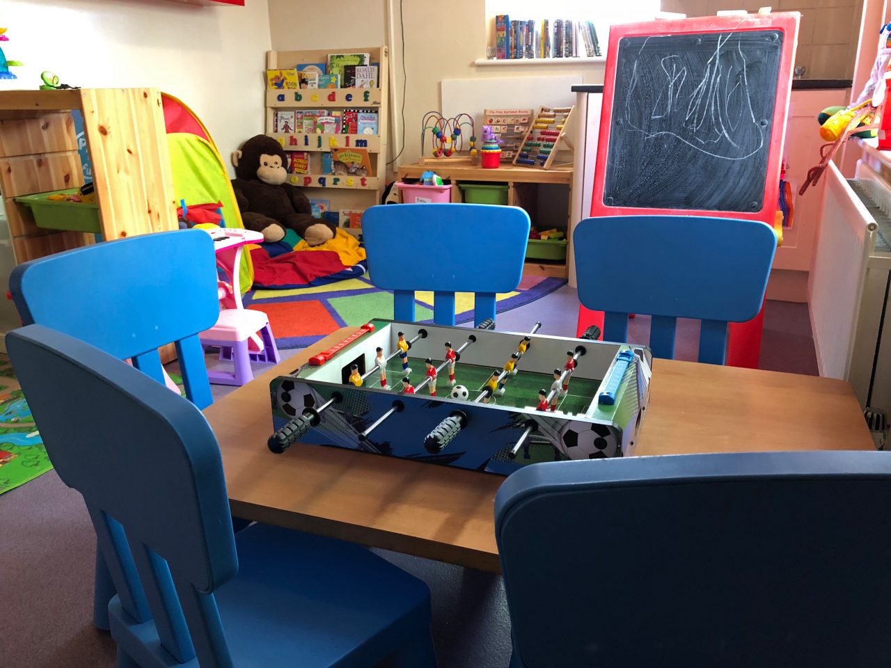 Children's toy room with a table and five blue chairs. In the background are lots of colourful toys and a blackboard.