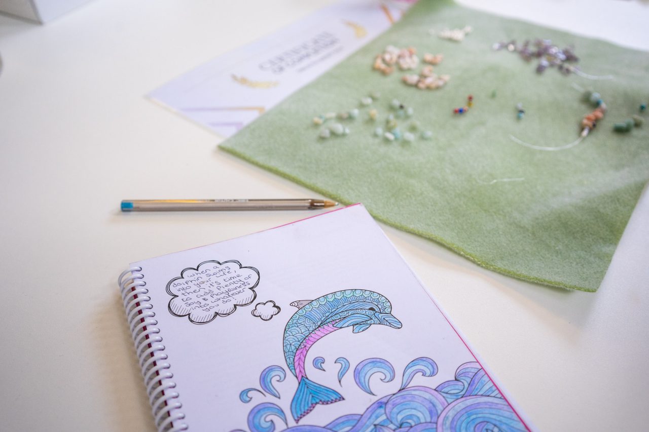 Open notebook of a drawing of a dolphin in the sea. Next to is a sheet of green paper with beads glued onto it.