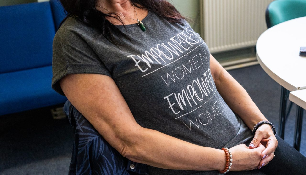 A person with shoulder-length hair sitting in a chair with their hands clasped in their lap. On their shirt is the phrase, 'Empowered women empower women'.