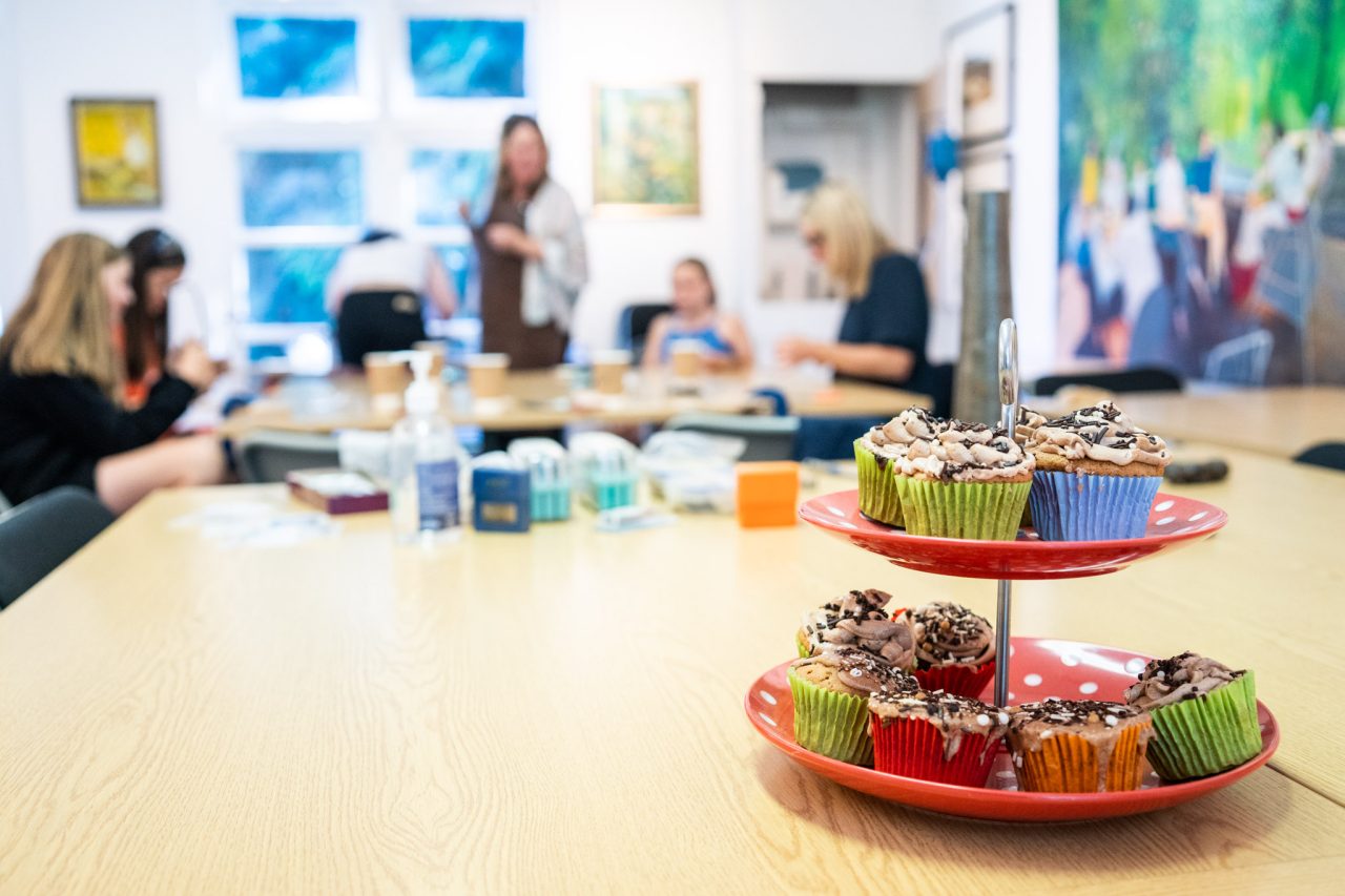 Cupcake stand, filled with cupcakes in colourful cases, sits on a table. Seven people are sitting in the background.