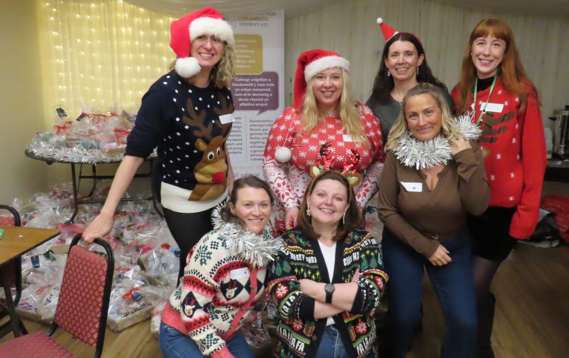 A group of people posing indoors at a festive event, wearing Christmas jumpers and Santa hats. Some are standing and others are seated, with wrapped gift packages arranged on tables behind them