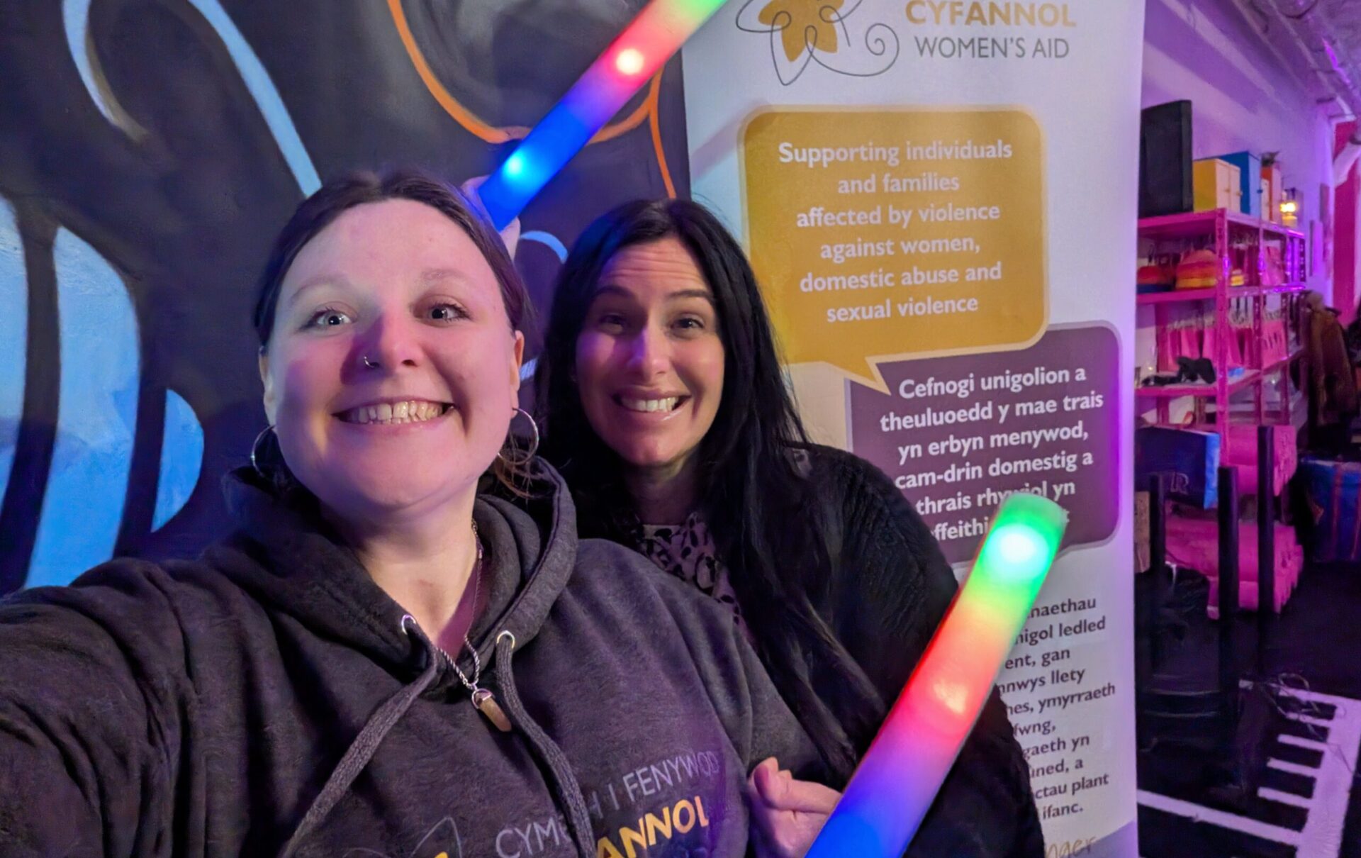 Two people standing close together indoors, facing the camera, holding colourful glow sticks. A Cyfannol Women’s Aid banner stands behind them with text about supporting individuals and families affected by domestic and sexual violence.