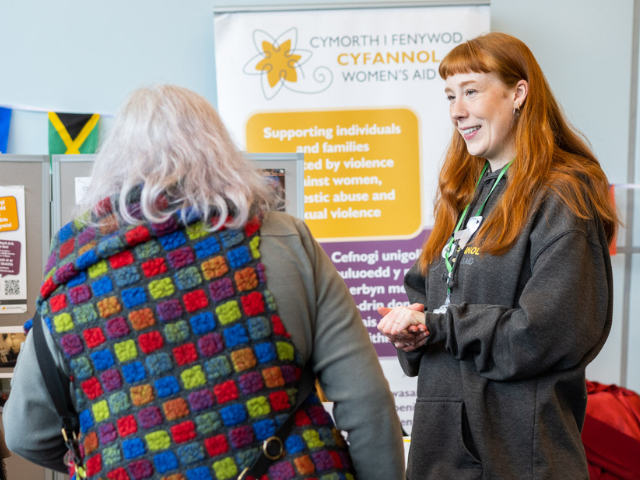Two people standing at an information stall. One person faces away wearing a multicoloured knitted vest, while another stands facing them in a dark hoodie and lanyard. Behind them is a banner reading ‘Cymorth i Fenywod Cyfannol Women’s Aid’ with text about supporting individuals and families affected by violence against women, domestic abuse and sexual violence.