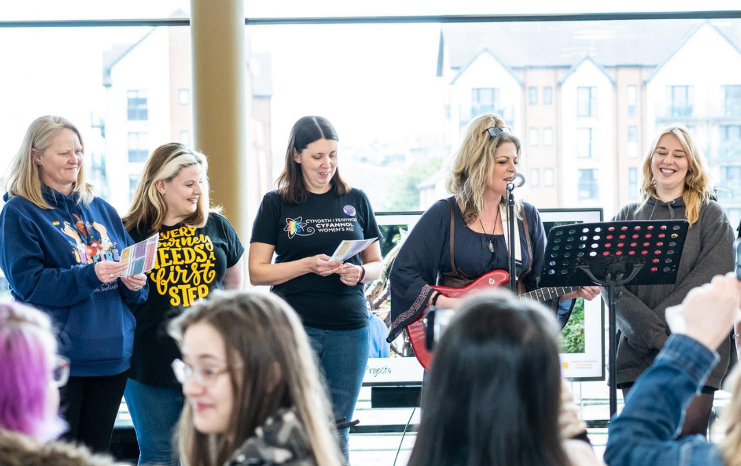 A small group of people standing indoors near large windows, holding printed sheets and facing a microphone and music stand. One person holds a guitar, and the group poised to begin singing.