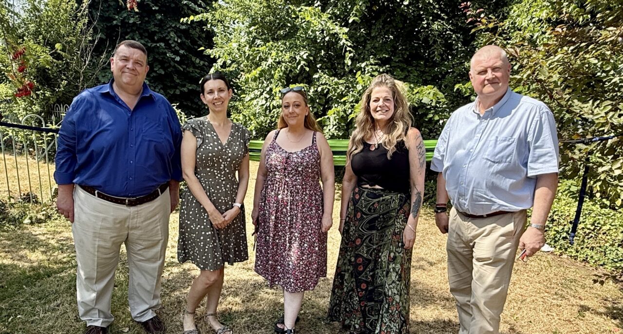 Two representatives from Monmouthshire Freemasons and three representatives from Cyfannol Women's Aid stand together in a woodland area behind a fire pit and in front of a green hammock during a visit to learn more about project.