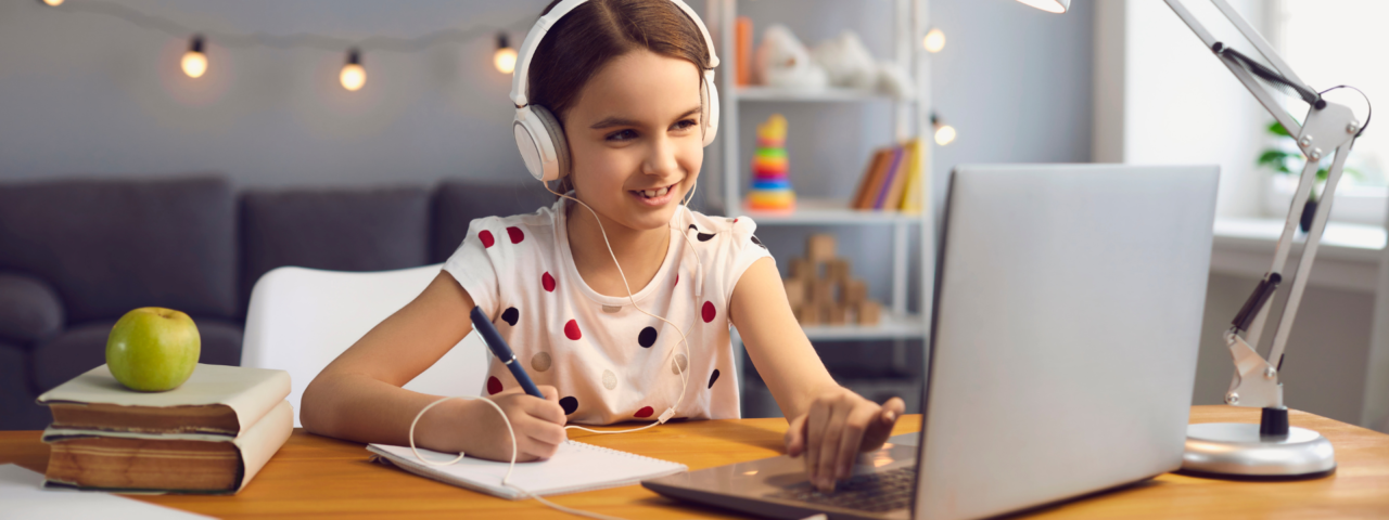 Child with brown, wearing headphone looking at a laptop screen with a pen and notepad in bedroom.