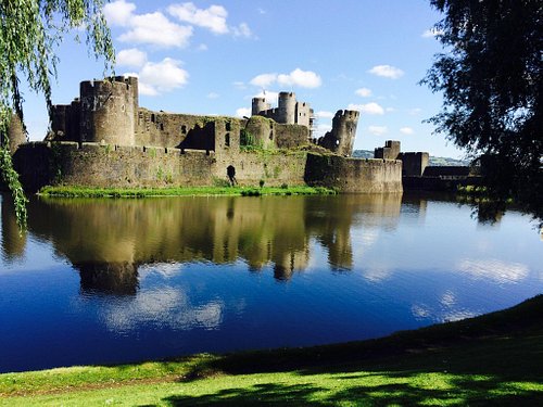 Image of Caerphilly castle on a sunny day with a few clouds in the sky and the reflection of the castle in the water.